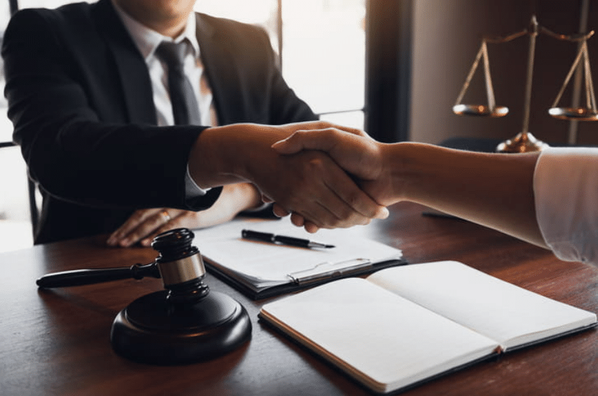 laywer shaking hands with a client. There's an open notebook between them, along with a clipboard and papers and a gavel.