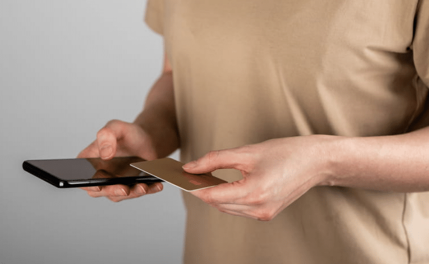 A person wearing a tan shirt. The photo is a close up of their hands holding a credit card and smartphone to pay off credit card.