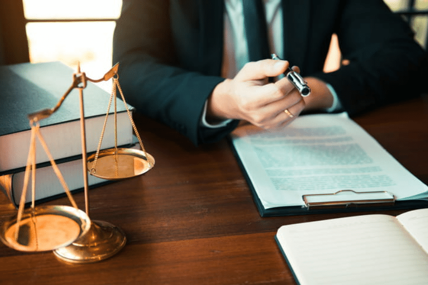 a lawyer holding out a pen for their client to sign paperwork regarding Chapter 7 bankruptcy. On the desk is a clipboard with papers, a stack of books and the scales of justice.